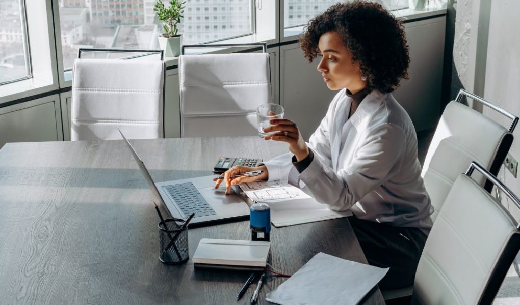 A woman sitting on a large desk, a laptop and papers on the desk, she is holding a glass -how to attract aligned clients tips