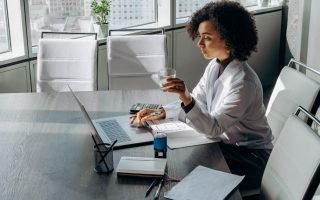 A woman sitting on a large desk, a laptop and papers on the desk, she is holding a glass -how to attract aligned clients tips